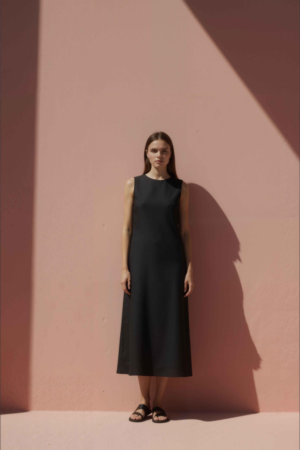 Woman in a black cotton shift dress standing against a pink wall.