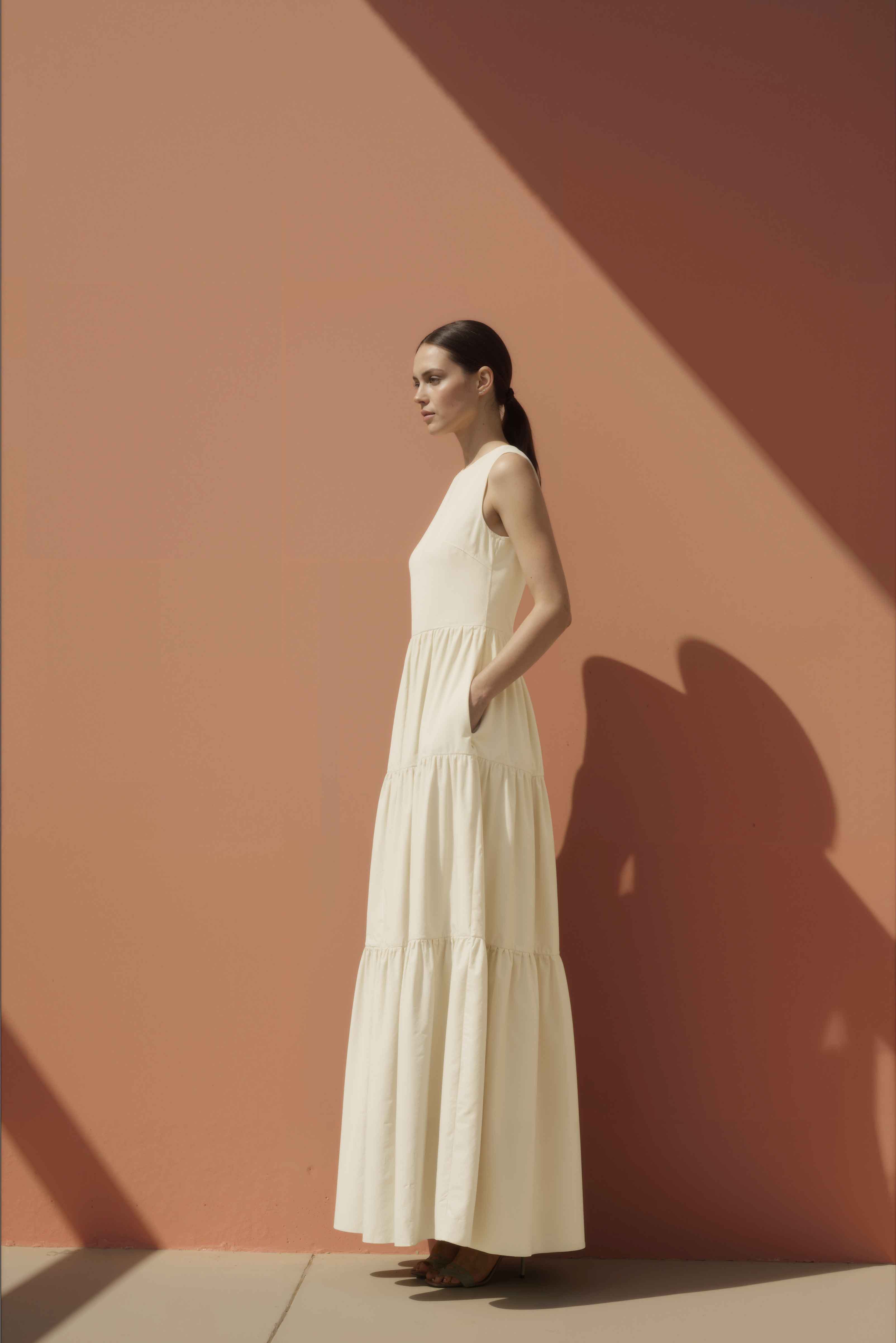 Woman in a white cotton maxi dress standing against a pink wall.