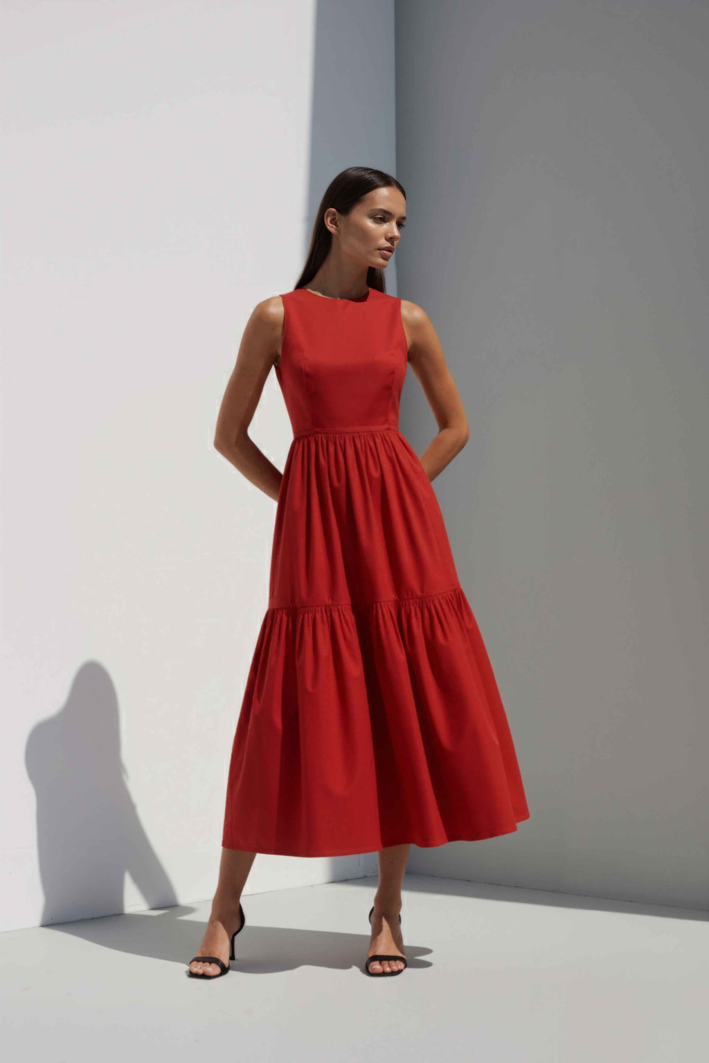 Anasua The Forme Edit Poplin Cotton Midi Dress in Sienna Red, a structured sleeveless cotton midi dress with a tiered skirt, photographed under natural sunlight.